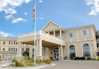 Exterior view of a large, beige senior living facility building with a covered entrance, multiple windows, and an American flag flying on a flagpole in front. The sky is partly cloudy.