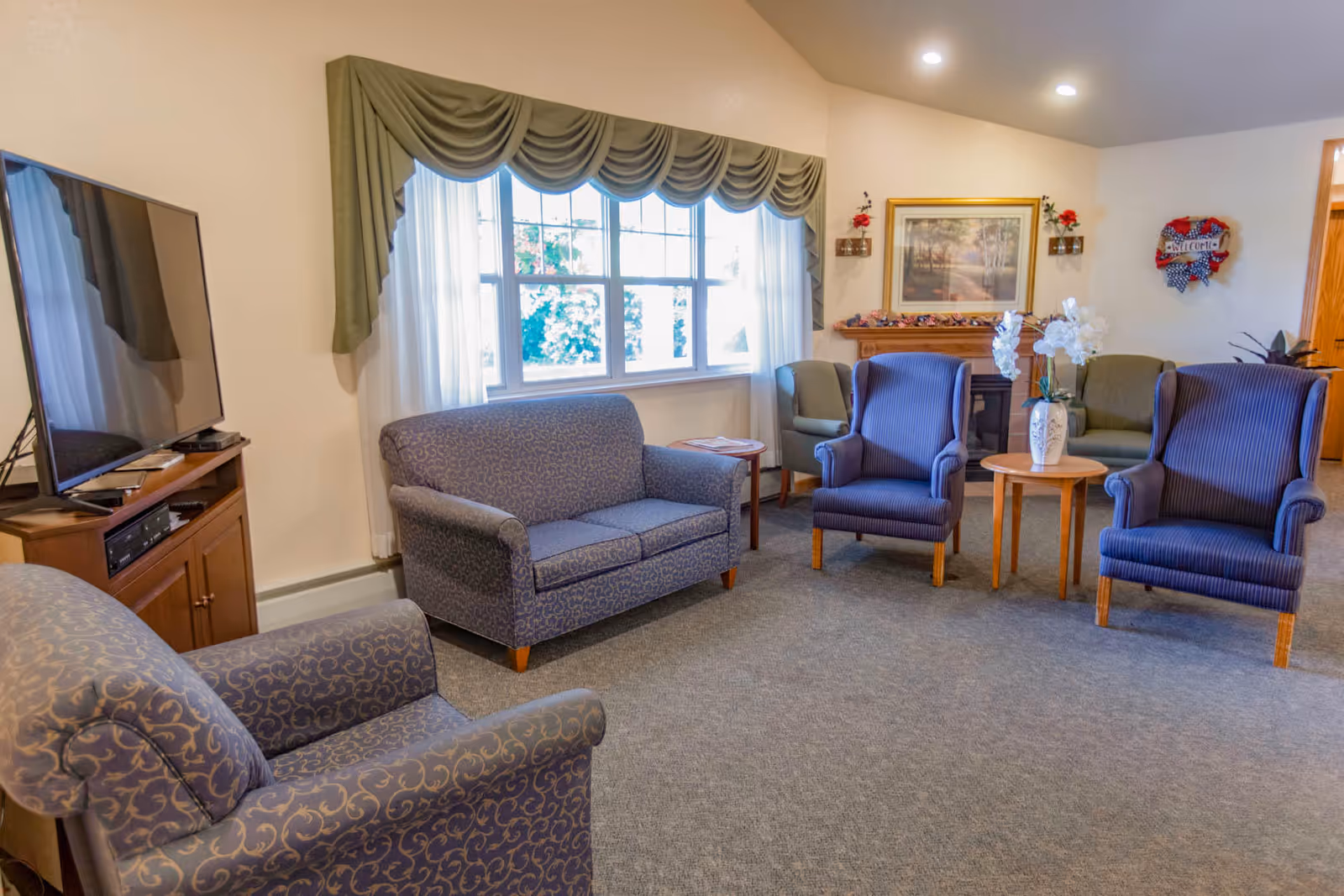 A cozy living room with patterned armchairs and a loveseat arranged around a small wooden table with a vase of white flowers. A large window with green valance and white curtains lets in natural light. A flat-screen TV sits on a wooden cabinet to the left. The room has a carpeted floor, a fireplace with decorative items, and framed artwork on the wall.
