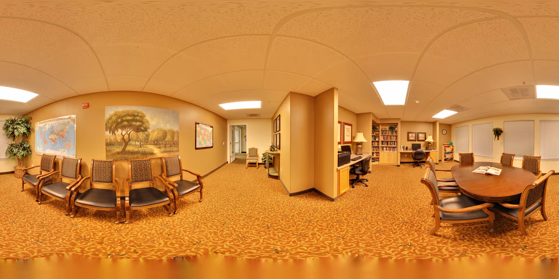A warm-toned senior living facility interior featuring a waiting area with wooden chairs lined up against the wall, decorated with a large painting of a tree and a world map. Adjacent to the waiting area is a workspace with desks, chairs, bookshelves, and lamps. On the right side, there is a conference or dining table with chairs around it, and windows with blinds. The room has patterned carpet flooring and a drop ceiling with fluorescent lighting.