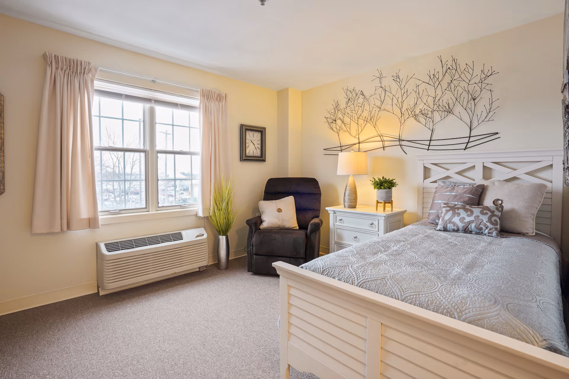 A cozy bedroom with a single bed featuring a white wooden frame and patterned bedding. Next to the bed is a white nightstand with a lamp and a small potted plant. A dark upholstered armchair with a cushion is placed near a window with beige curtains. The wall above the bed has a decorative metal art piece resembling tree branches, and a clock is mounted on the adjacent wall. The room has beige walls and carpeted flooring.