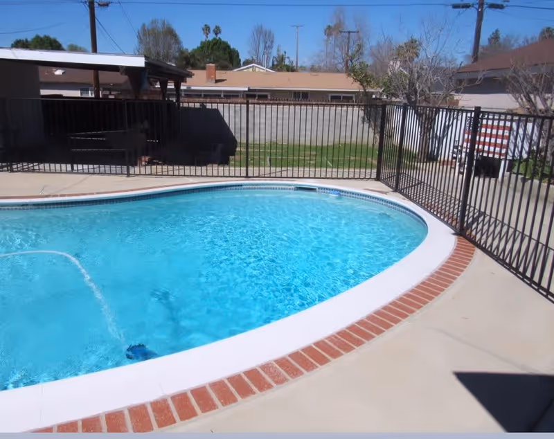 Outdoor swimming pool with clear blue water surrounded by a concrete deck and a black metal safety fence. In the background, there are residential houses, trees, and a clear blue sky.