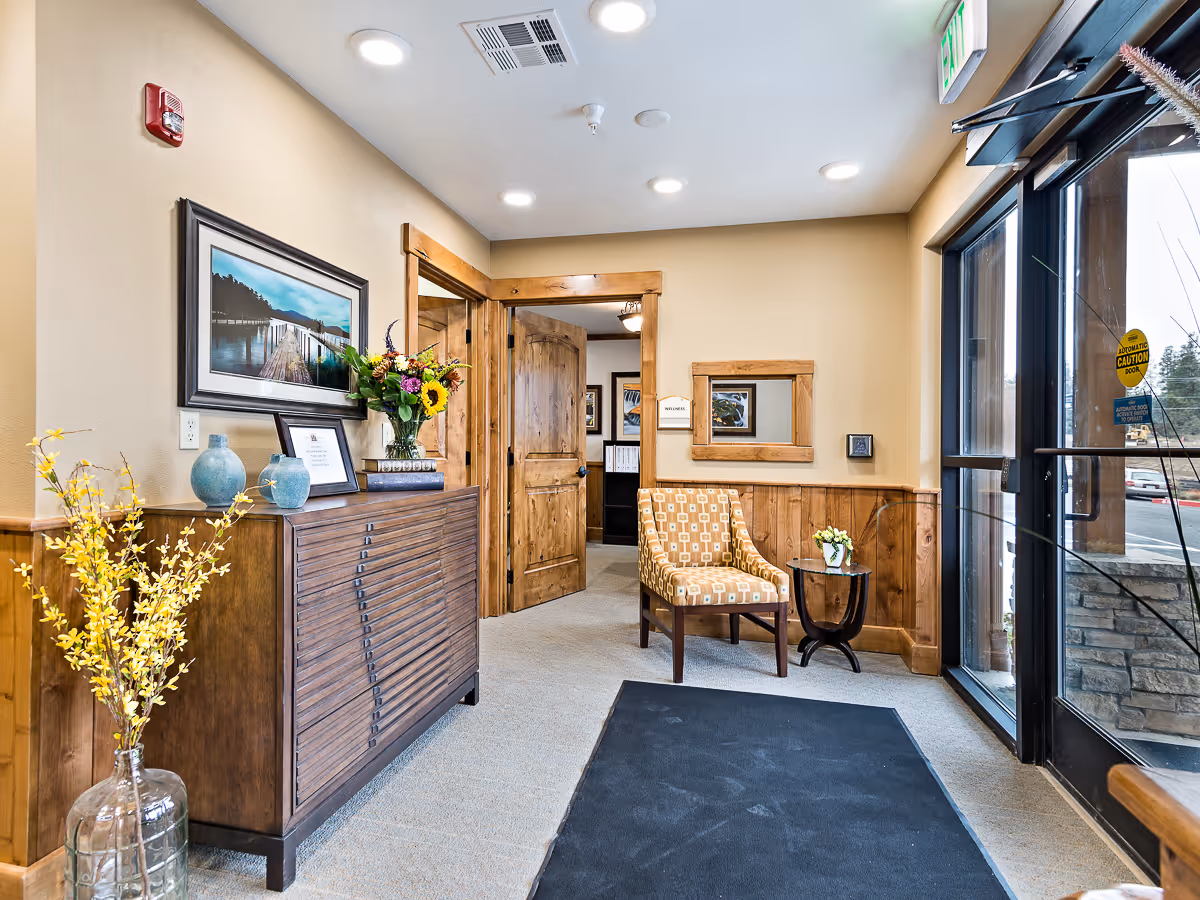 Entrance area of Mill View Memory Care facility with a wooden cabinet decorated with vases and flowers on the left, a patterned armchair next to a small round table with a flower pot on the right, beige walls with wooden paneling, a framed picture on the wall, and glass doors leading outside.