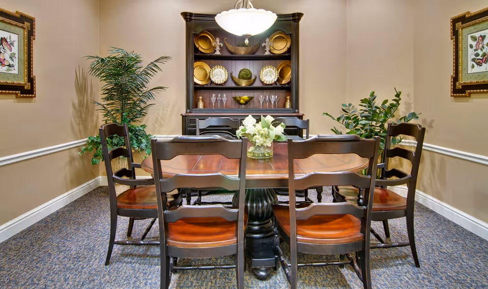 A dining room with a wooden dining table surrounded by six wooden chairs. The table has a glass vase with white flowers in the center. Behind the table is a dark wooden hutch displaying decorative plates, bowls, and glassware. The room has beige walls with white trim, two framed butterfly pictures on either side, and two green potted plants in the corners. A ceiling light fixture hangs above the table.
