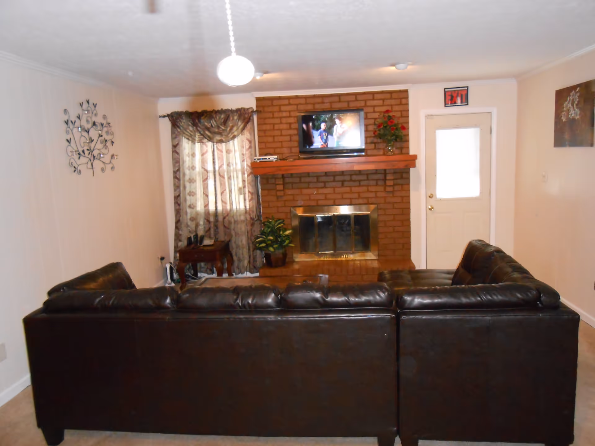 A living room with a large dark brown leather sectional sofa facing a brick fireplace. Above the fireplace is a mounted flat-screen TV showing a scene with people. To the left of the fireplace is a window with patterned curtains and a small wooden side table with a phone. There is a potted plant on the hearth. To the right of the fireplace is a white door with an exit sign above it. The walls are light-colored with decorative wall art on both sides.