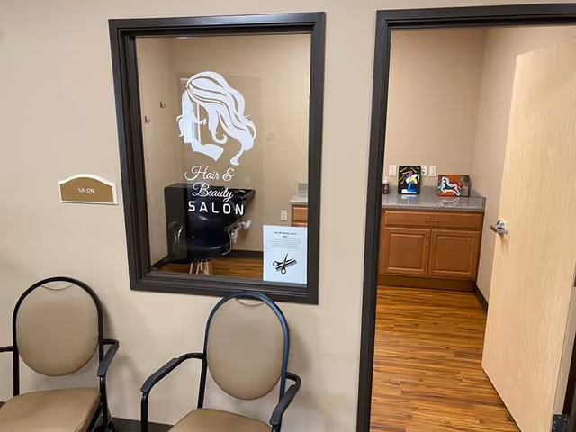 Interior view of a hair and beauty salon in an assisted living facility. The image shows a window with a salon logo featuring a woman's silhouette and the text 'Hair & Beauty SALON'. Two beige chairs with black armrests are placed against the wall outside the salon. Inside the salon, a black salon chair and a countertop with cabinets are visible. The floor has a wood finish.