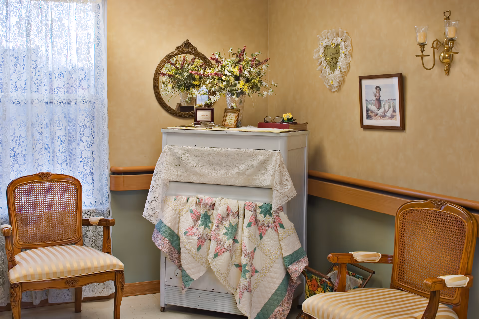 A cozy corner in a senior living facility with two wooden chairs featuring cane backs and striped cushions. Between the chairs is a small white cabinet draped with a lace cloth and a colorful quilt. On top of the cabinet are framed pictures, a vase with a floral arrangement, and a pair of glasses. The wall behind has a round mirror, a heart-shaped decoration, a framed picture, and a brass wall sconce with two candle-style lights. A window with lace curtains is on the left side.