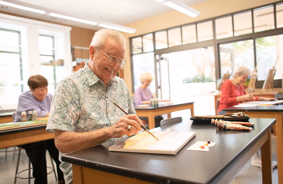 A group of elderly people sitting at tables in a bright room, engaged in painting on canvases. The focus is on an elderly man smiling as he paints, with other seniors painting in the background.