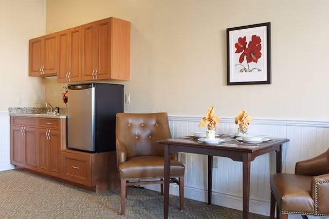 A small dining area in an assisted living facility featuring a wooden table set for two with folded napkins, two brown leather chairs, a kitchenette with wooden cabinets, a small refrigerator, and a framed floral artwork on the wall.