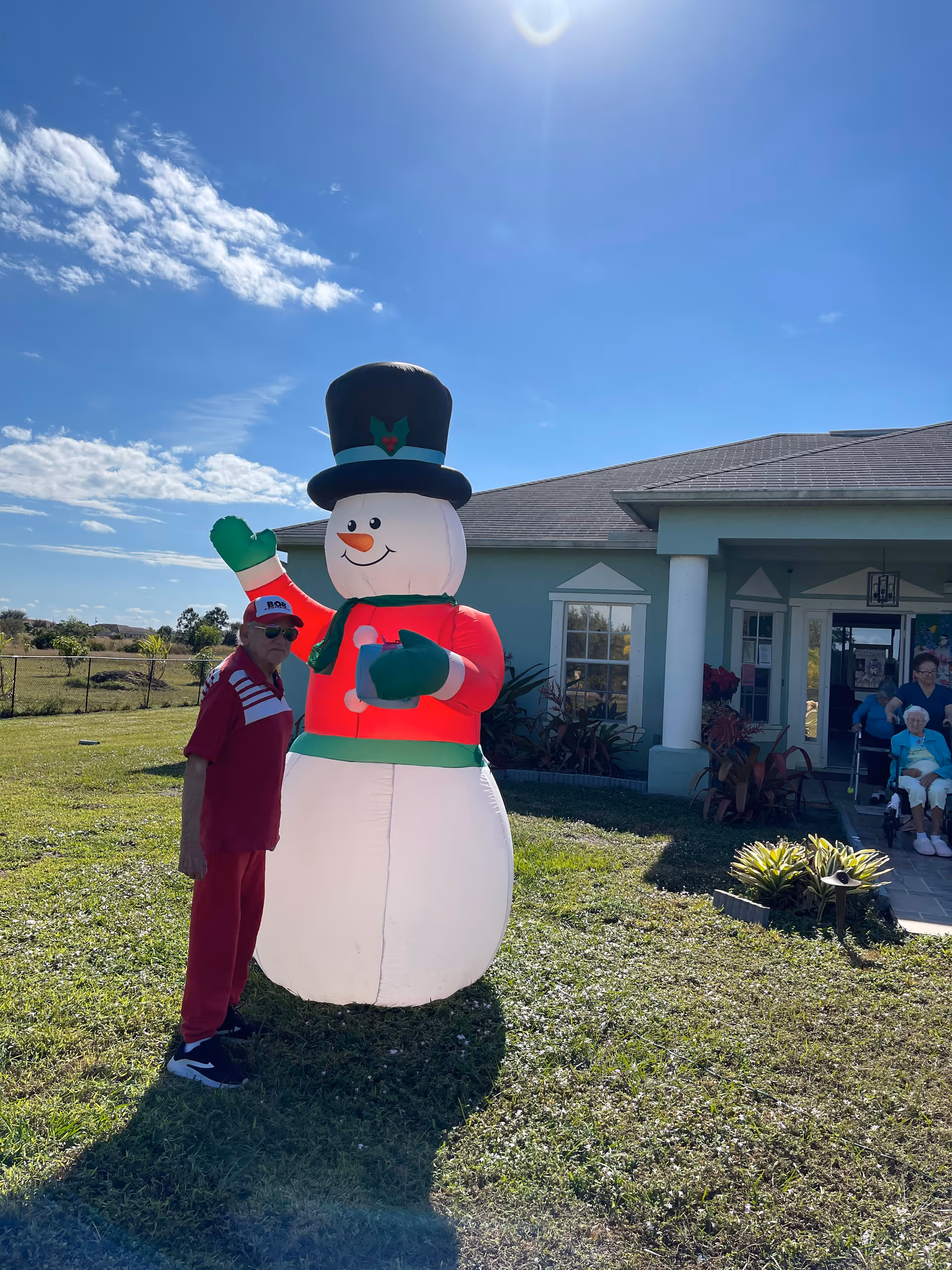 An elderly man dressed in red stands next to a large inflatable snowman decoration on a grassy lawn outside a single-story building. In the background, two elderly women are seated near the entrance of the building under a covered porch. The sky is clear and sunny.