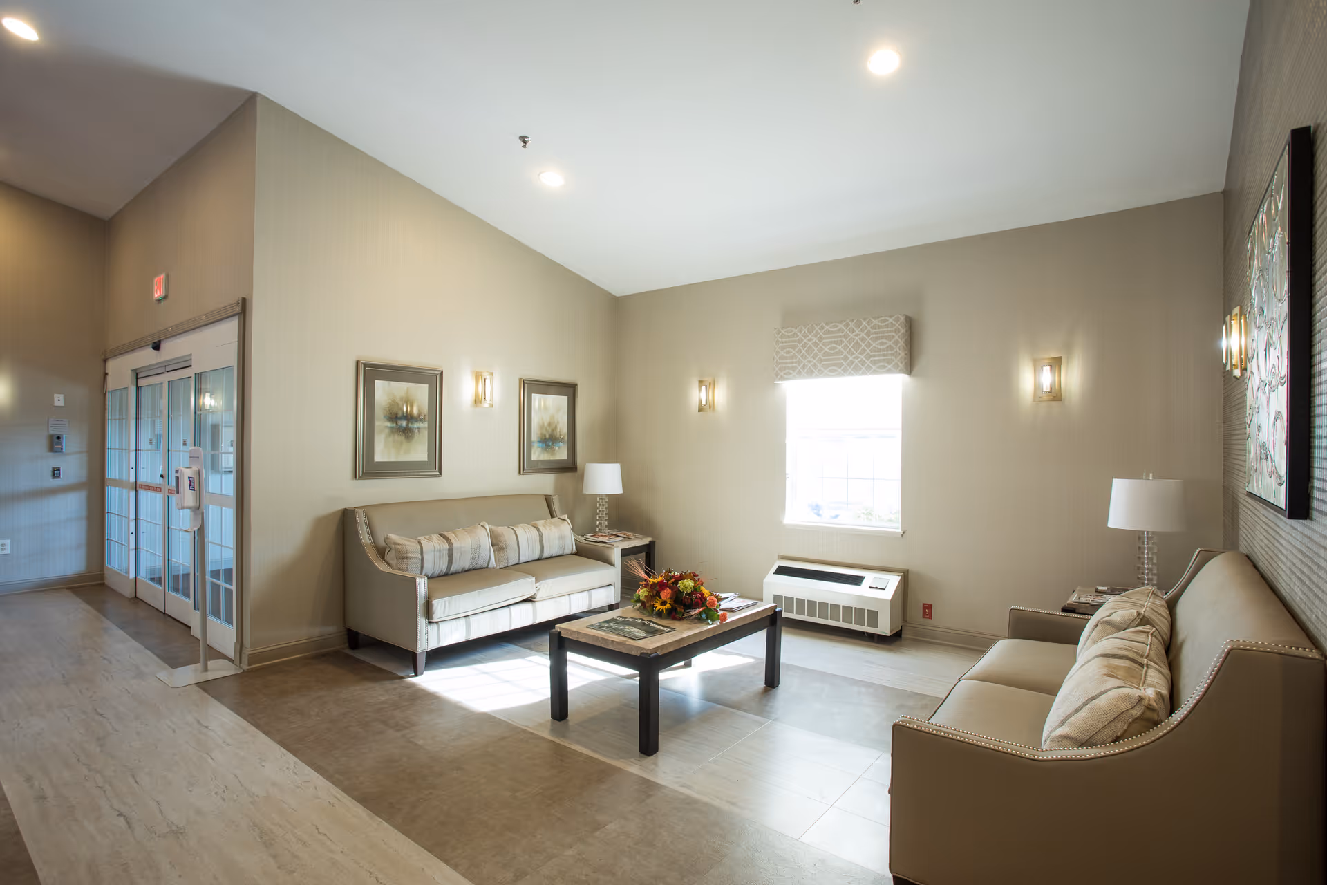 A bright and clean living room area in a senior living facility with two beige sofas facing each other, a coffee table with a floral centerpiece in the middle, wall-mounted lights, framed artwork, a window with a patterned valance, and a glass double door entrance.