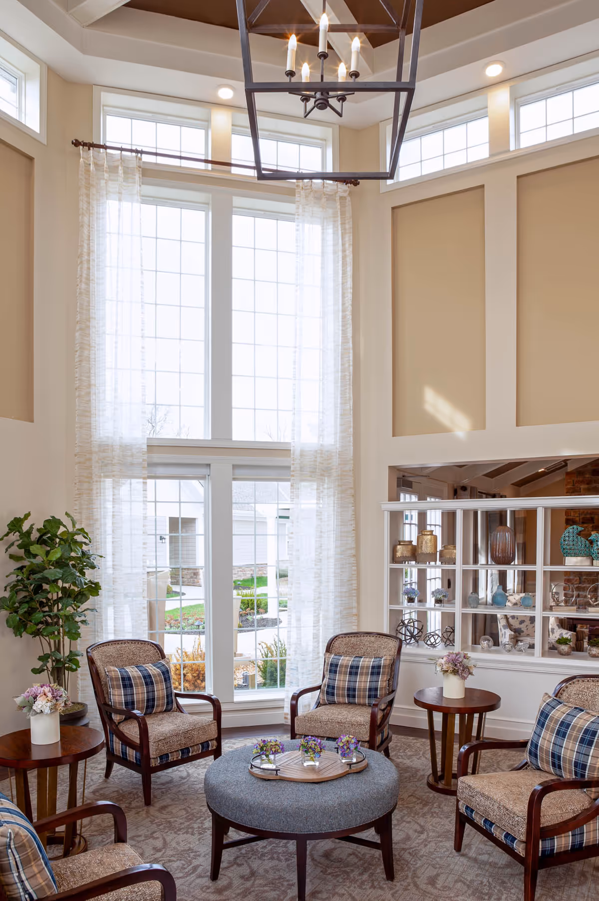 A bright and cozy living room area with four cushioned armchairs featuring plaid pillows arranged around a round ottoman with a wooden tray holding small flower arrangements. The room has tall windows with sheer curtains, beige walls with white trim, a high ceiling with a modern chandelier, and decorative shelving with vases and ornaments.