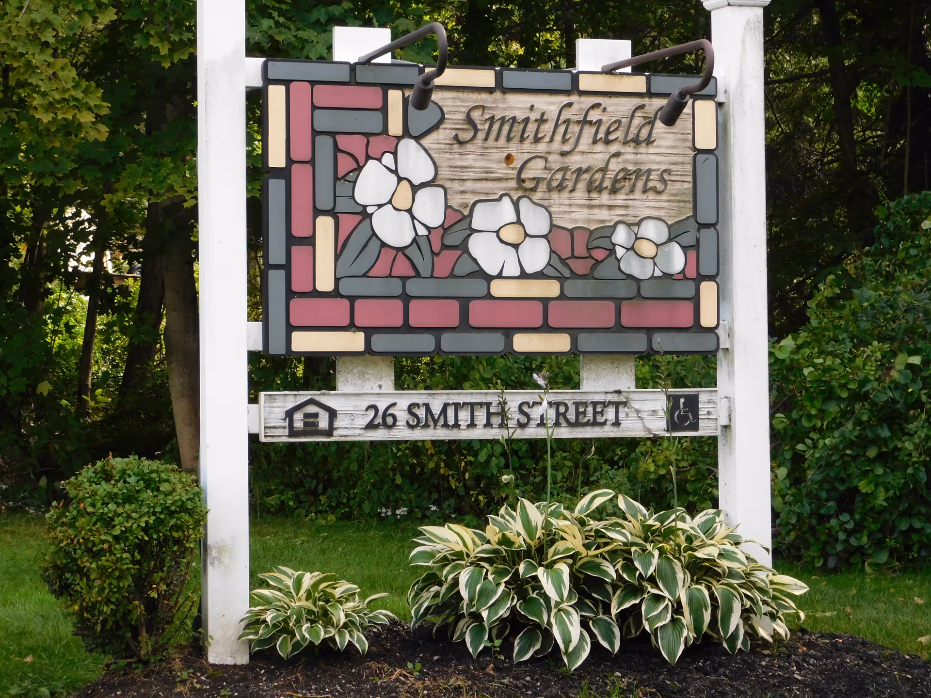 A decorative wooden sign for Smithfield Gardens featuring a floral design with white flowers and green leaves, mounted on white posts surrounded by green bushes and plants.