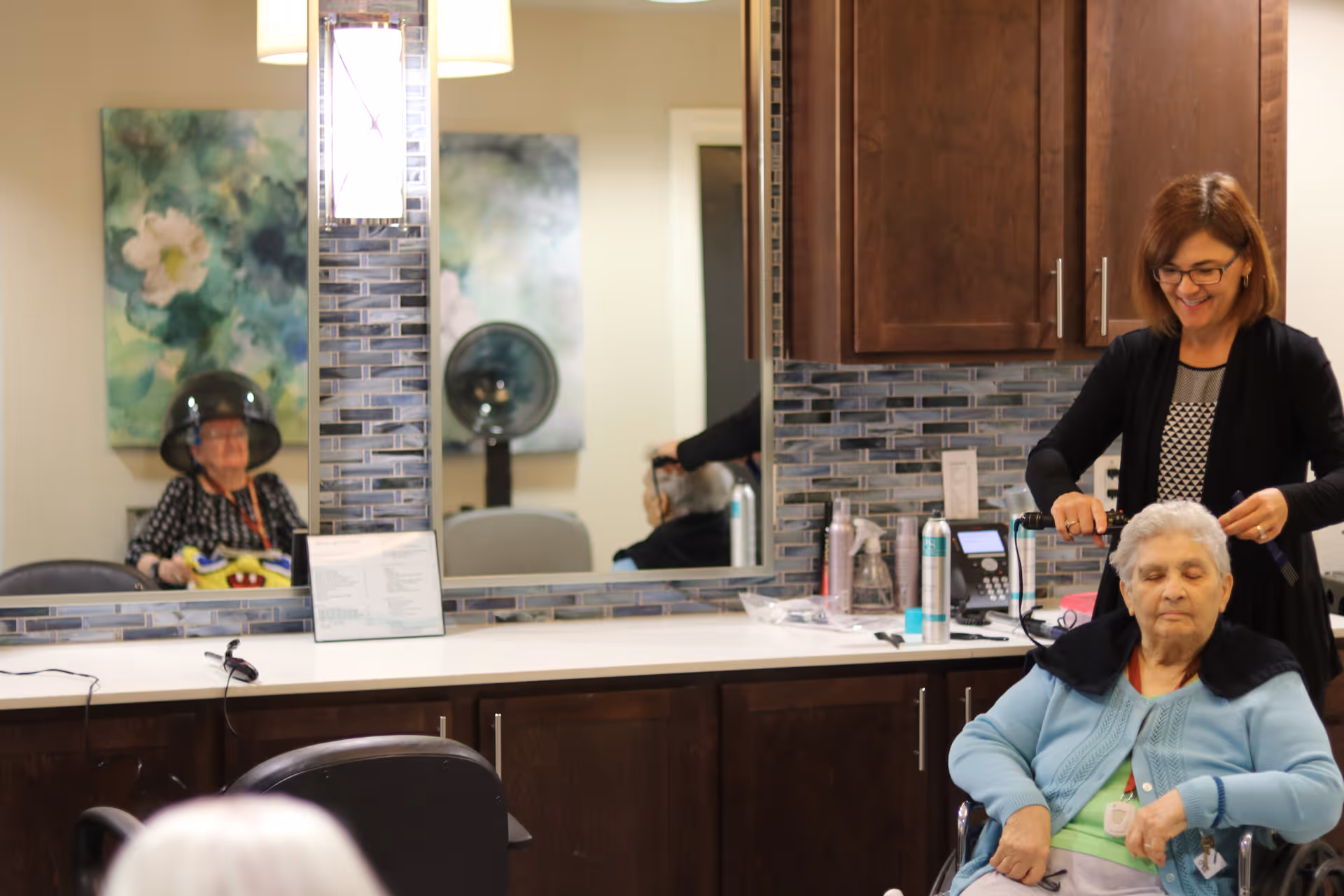 A hair salon area inside an assisted living facility where a woman is styling the hair of an elderly woman seated in a wheelchair. Another elderly woman is visible in the mirror reflection sitting under a hair dryer. The room has dark wood cabinets, a tiled backsplash, and a large mirror.