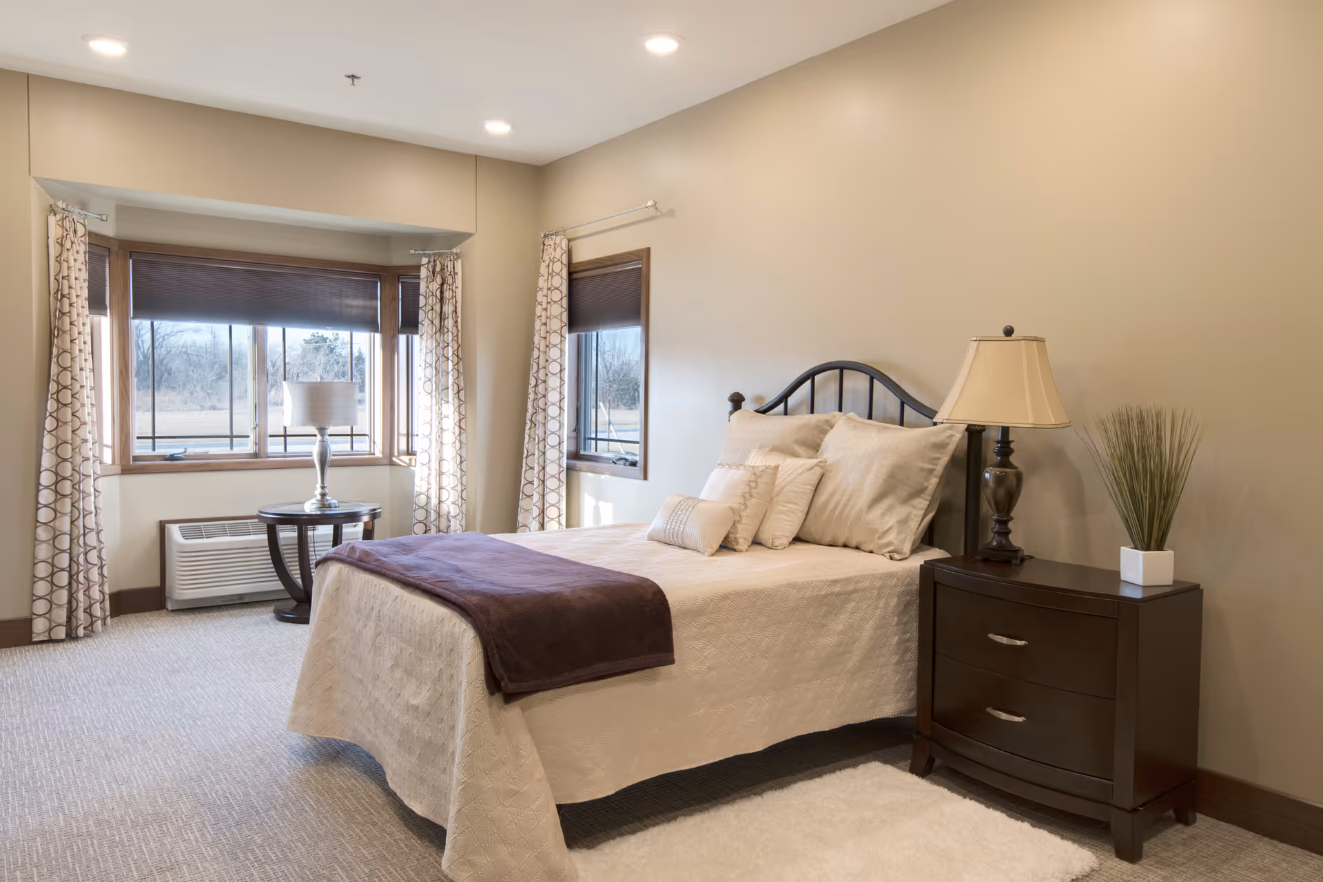 A cozy bedroom with a neatly made bed featuring beige bedding and multiple pillows. There is a dark wooden nightstand with a lamp and a small plant on it. The room has beige walls, carpeted floor, and a large window with patterned curtains letting in natural light. A small round table with a lamp is placed near the window.
