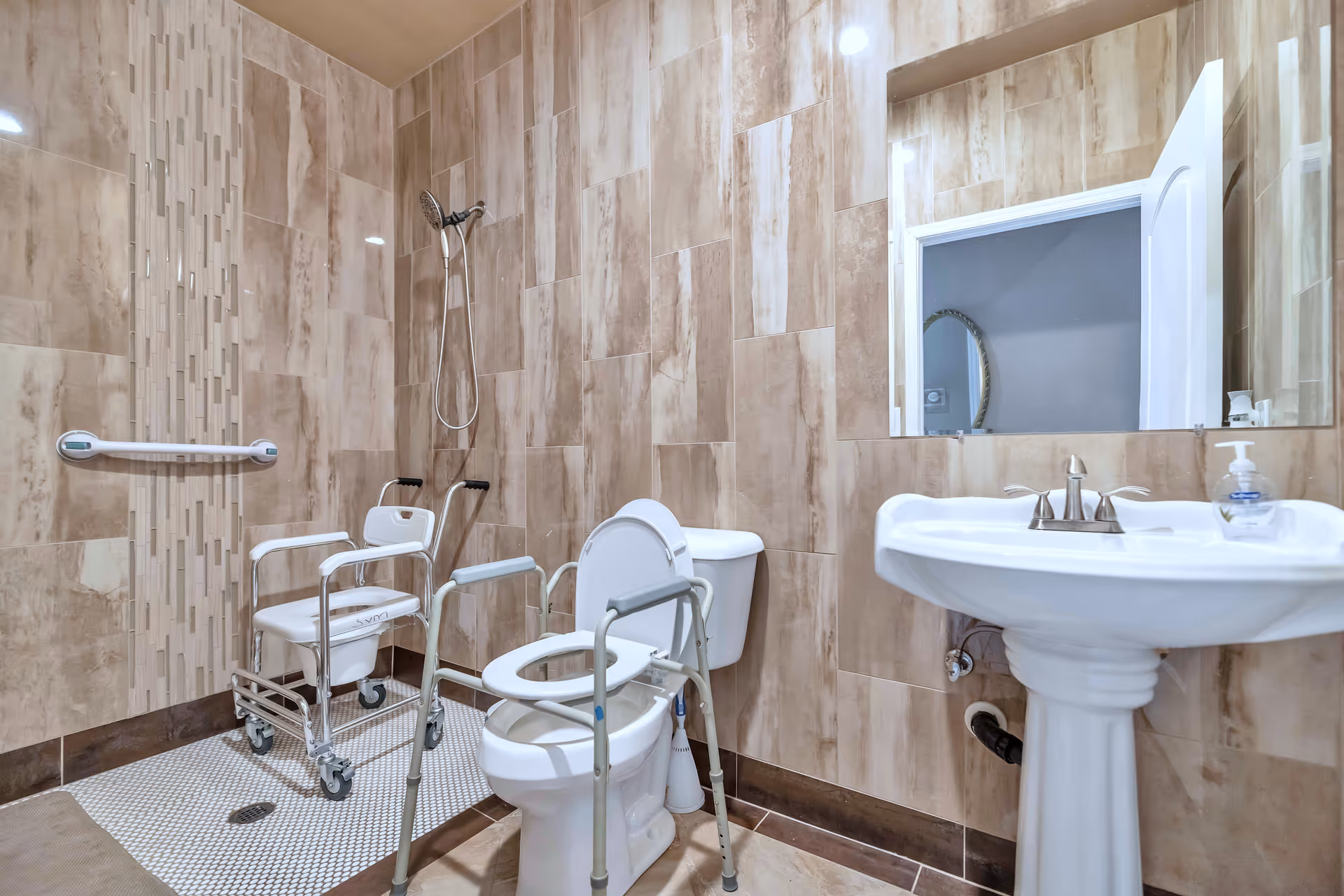 Accessible tiled bathroom featuring a roll-in shower with a shower chair, grab bars, a raised toilet with support rails, and a pedestal sink.