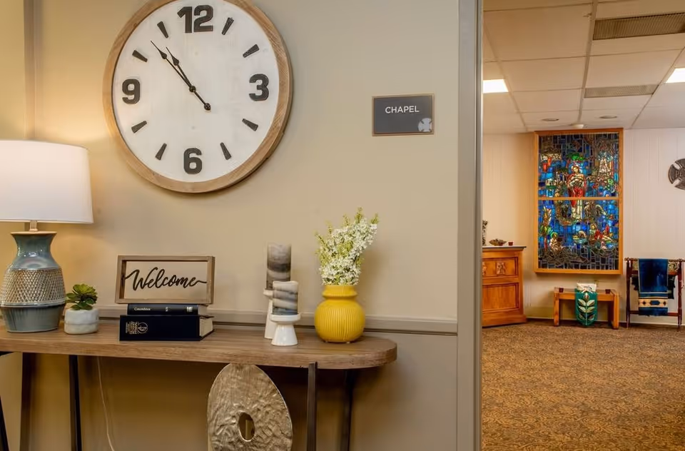 Interior view of a hallway corner in a senior living facility with a wooden table holding a lamp, a small plant, a 'Welcome' sign, books, decorative items, and a yellow vase with white flowers. A large round wall clock shows the time as 11:55. A sign on the wall reads 'CHAPEL'. In the background, there is a room with a stained glass window and wooden furniture.
