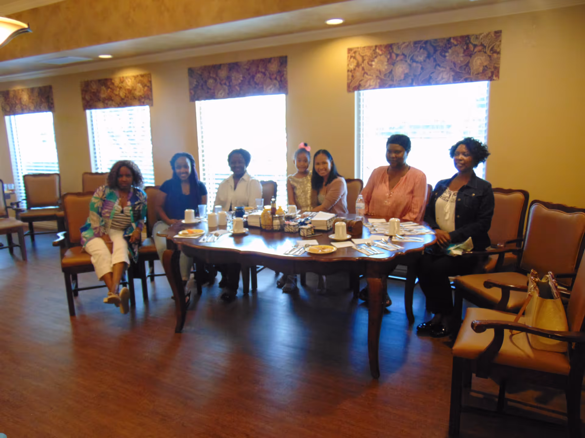A group of six women and one young girl sitting around a wooden dining table set with plates, cups, and utensils in a well-lit room with large windows and floral valances.