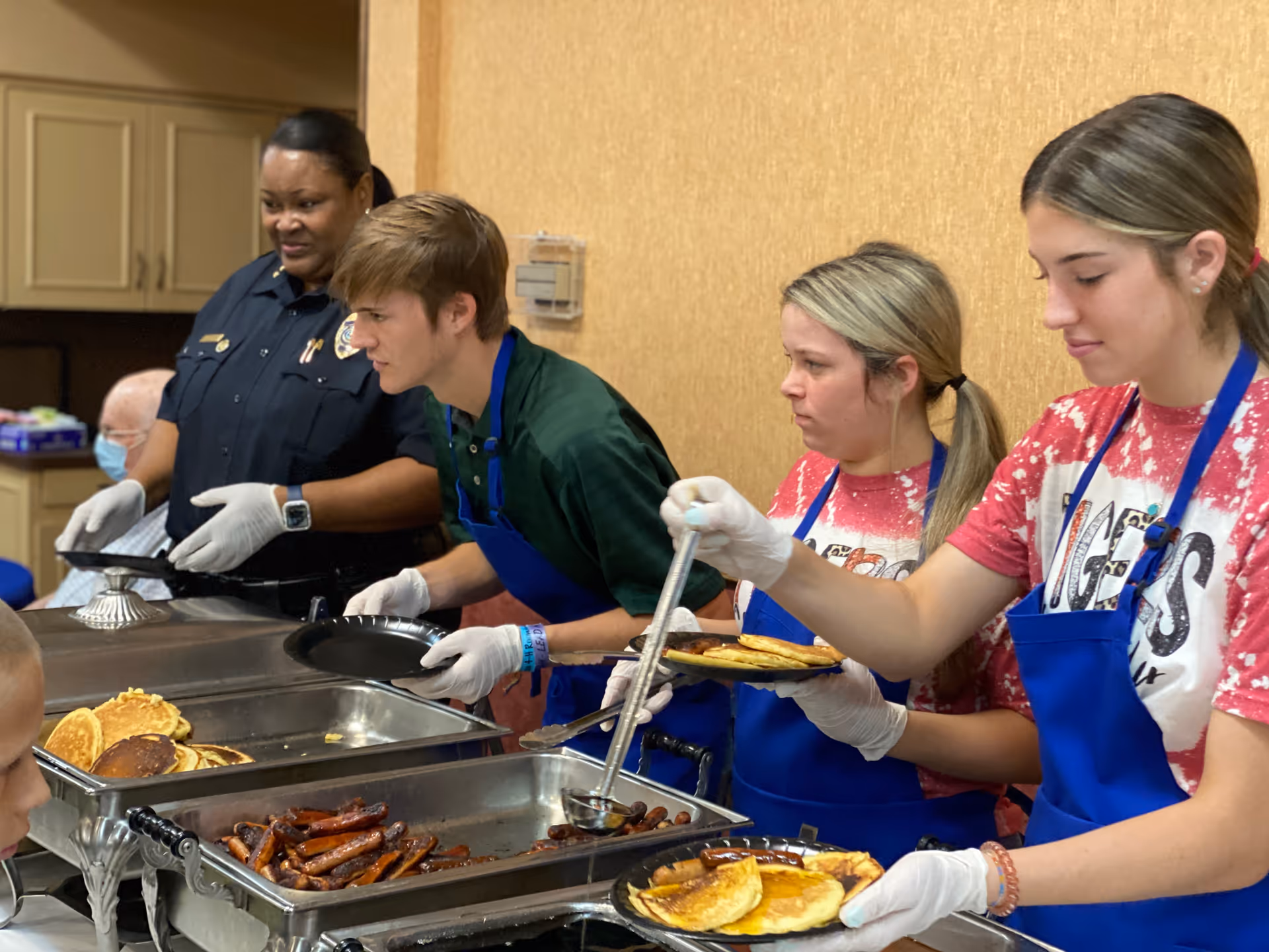 Three young volunteers wearing blue aprons and gloves serve pancakes and sausages from buffet trays to seated elderly individuals in a dining area. A woman in a dark uniform stands nearby assisting with the serving.