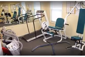 A small fitness room with exercise equipment including a treadmill, stationary bike, weight machine, and other workout apparatus. The room has beige walls, a large mirror on one side, and two windows letting in natural light.
