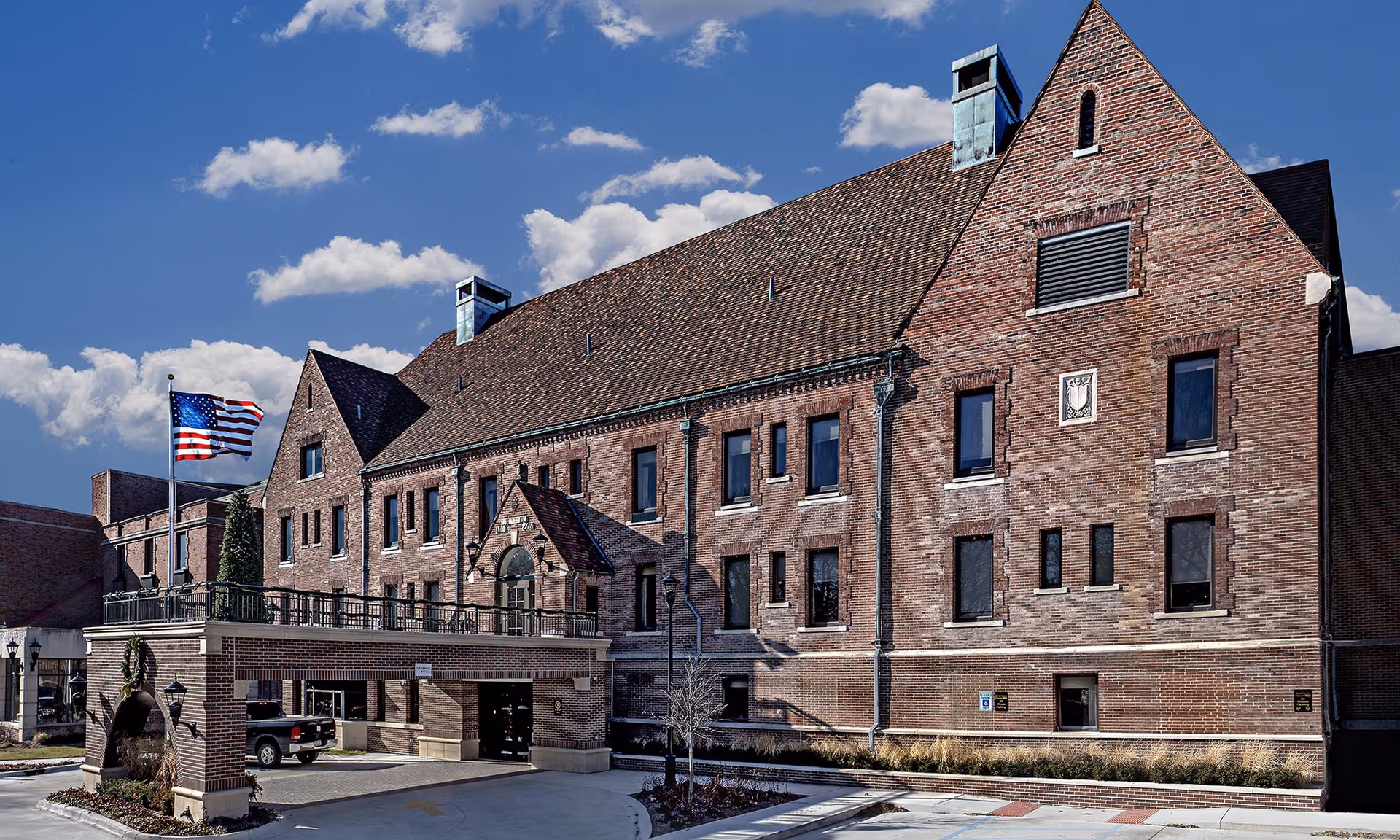 Exterior view of a large brick building with a steep roof under a blue sky with scattered clouds. The building has multiple windows and an entrance covered by a brick archway. An American flag is flying on a flagpole near the entrance.