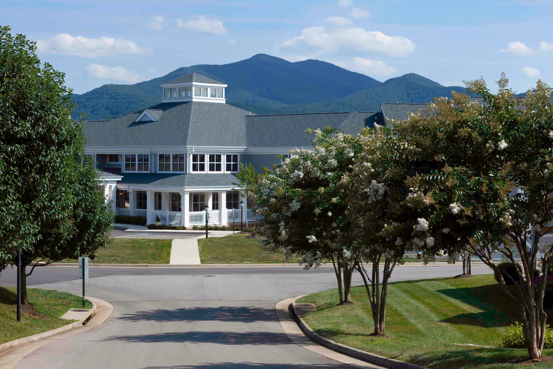 View of a large senior living facility building with a gray roof and white exterior, surrounded by green lawns and trees with white flowers, set against a backdrop of blue mountains under a partly cloudy sky.