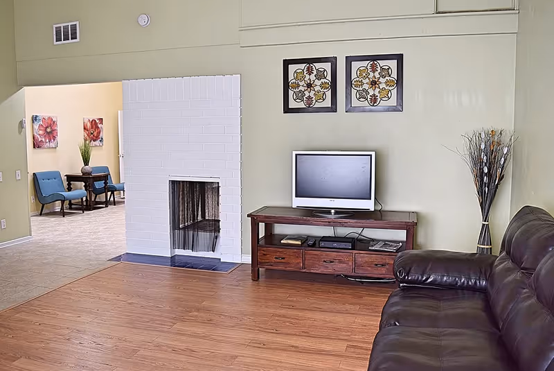 Living room with a leather sofa, television on a wooden console, a white brick fireplace, and a seating nook visible through an open doorway.