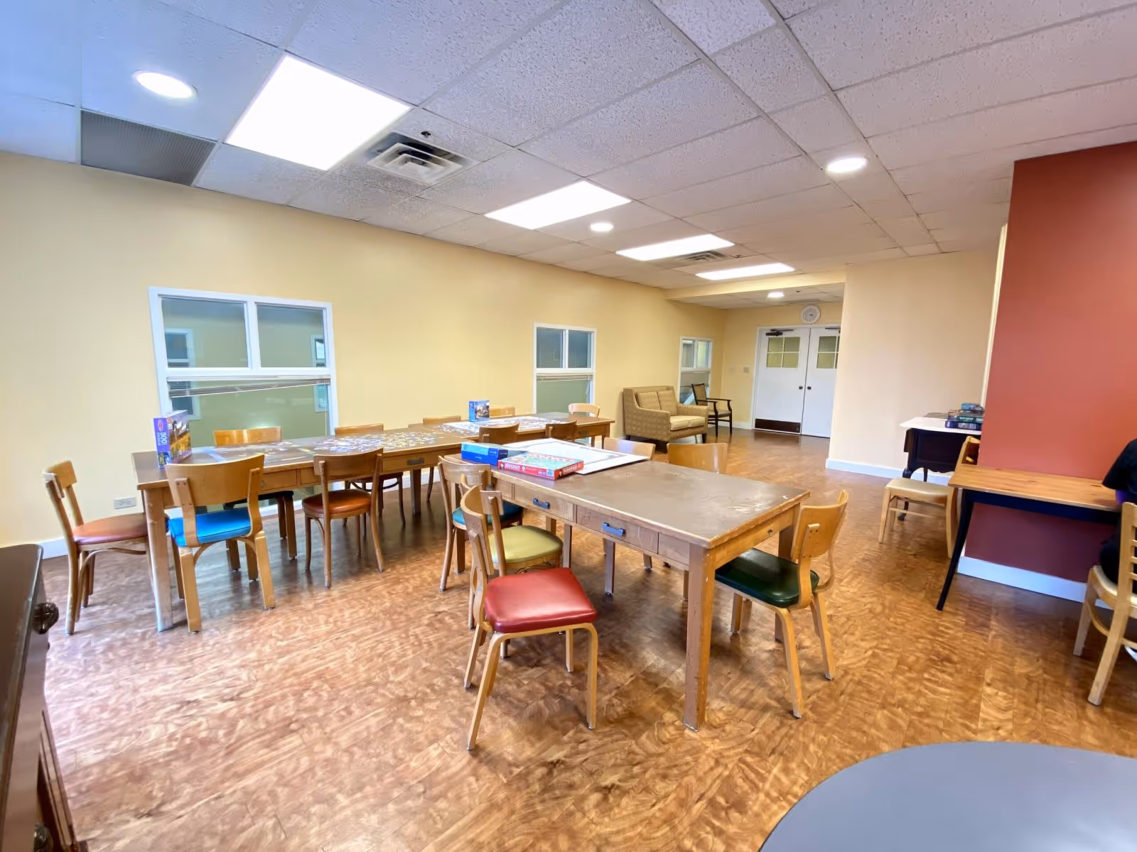 Bright common room with long wooden tables and mixed-color chairs set up for activities and games.