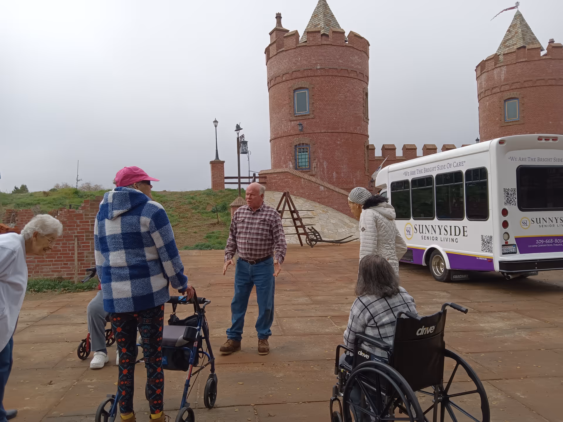 A group of elderly people gathered outdoors near a brick castle-like structure with two towers. One person is using a walker, another is in a wheelchair, and others are standing and conversing. A white and purple Sunnyside Senior Living shuttle bus is parked nearby.