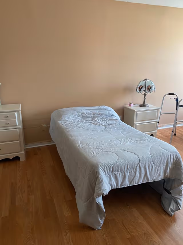 A simple bedroom with a single bed covered with a light gray quilt. There are two white nightstands on either side of the bed, one with a decorative lamp and a small item on top. The room has wooden flooring and beige walls. A walker is positioned near the right nightstand.