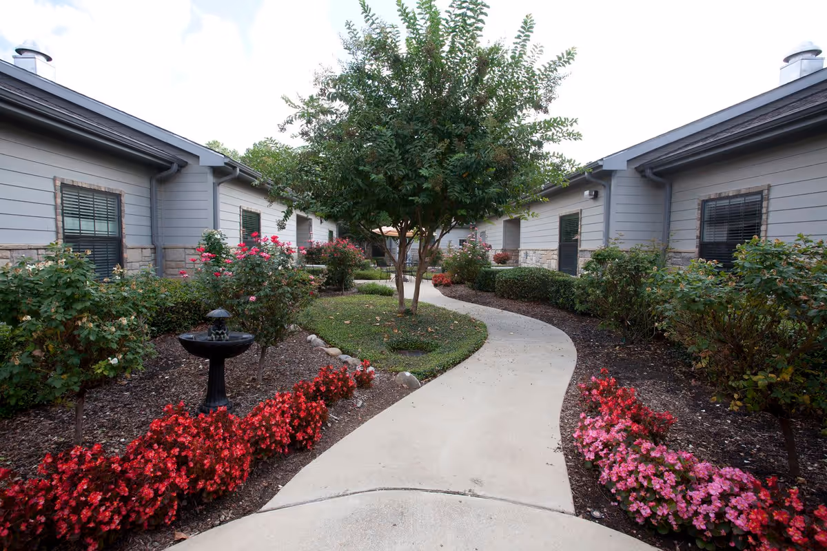 A landscaped outdoor courtyard with a winding concrete pathway flanked by vibrant red and pink flowers, bushes, and a small black birdbath. Two single-story buildings with gray siding and stone accents border the courtyard, and a tree stands in the center of the grassy area.