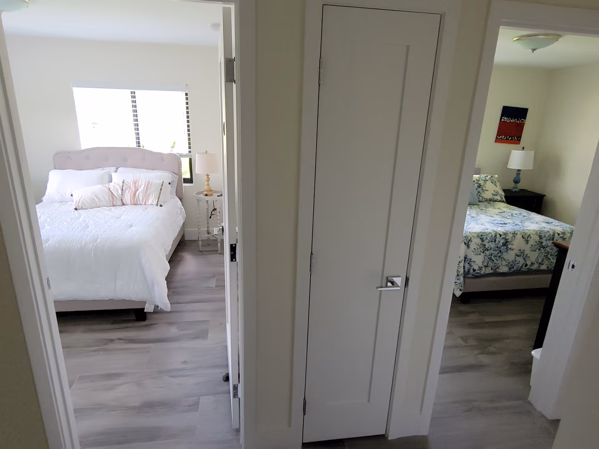 View of two bedrooms from a hallway in a senior living facility. The left bedroom has a white bed with multiple pillows and a bedside table with a lamp. The right bedroom has a floral-patterned bedspread, a bedside table with a lamp, and a piece of artwork on the wall. The floor is light-colored wood, and there is a closed white door in the center of the hallway.