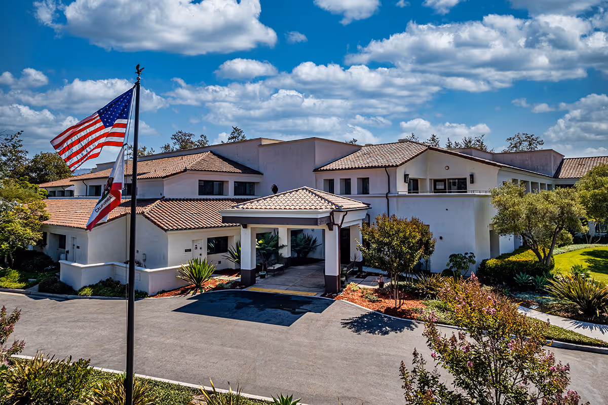 Exterior view of a senior living facility building with white walls and a tiled roof under a partly cloudy blue sky. There is a covered entrance with columns and landscaped greenery around the building. Two flagpoles display the American flag and the California state flag.