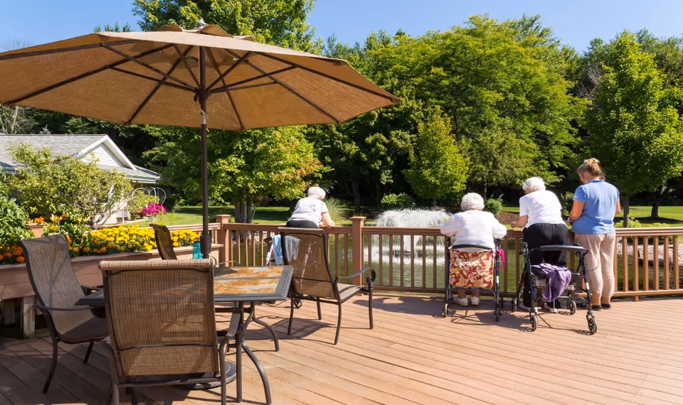 Sunny outdoor deck with patio furniture and a large umbrella overlooking a pond fountain where several people, some with walkers, stand at the railing.