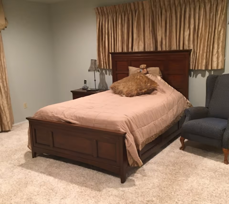 A cozy bedroom with a wooden bed frame, beige bedding, a brown decorative pillow, and a teddy bear on the bed. There is a wooden nightstand with a lamp and a phone, beige curtains covering the window, and a blue upholstered armchair on a light carpeted floor.