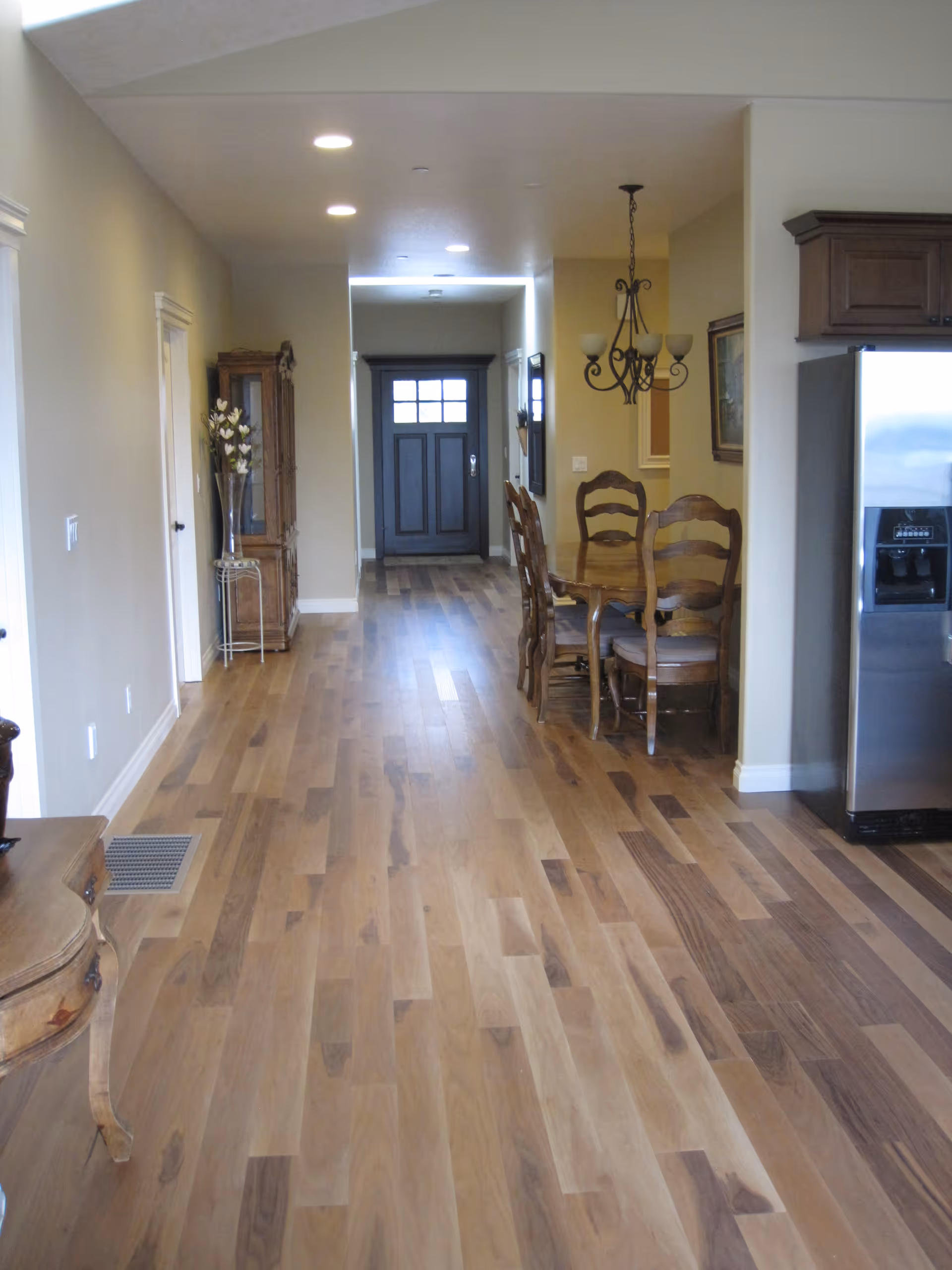 Interior view of a senior living facility hallway with wooden flooring, a dining table with chairs on the right side, a chandelier above the table, a stainless steel refrigerator, and a dark front door at the end of the hallway.