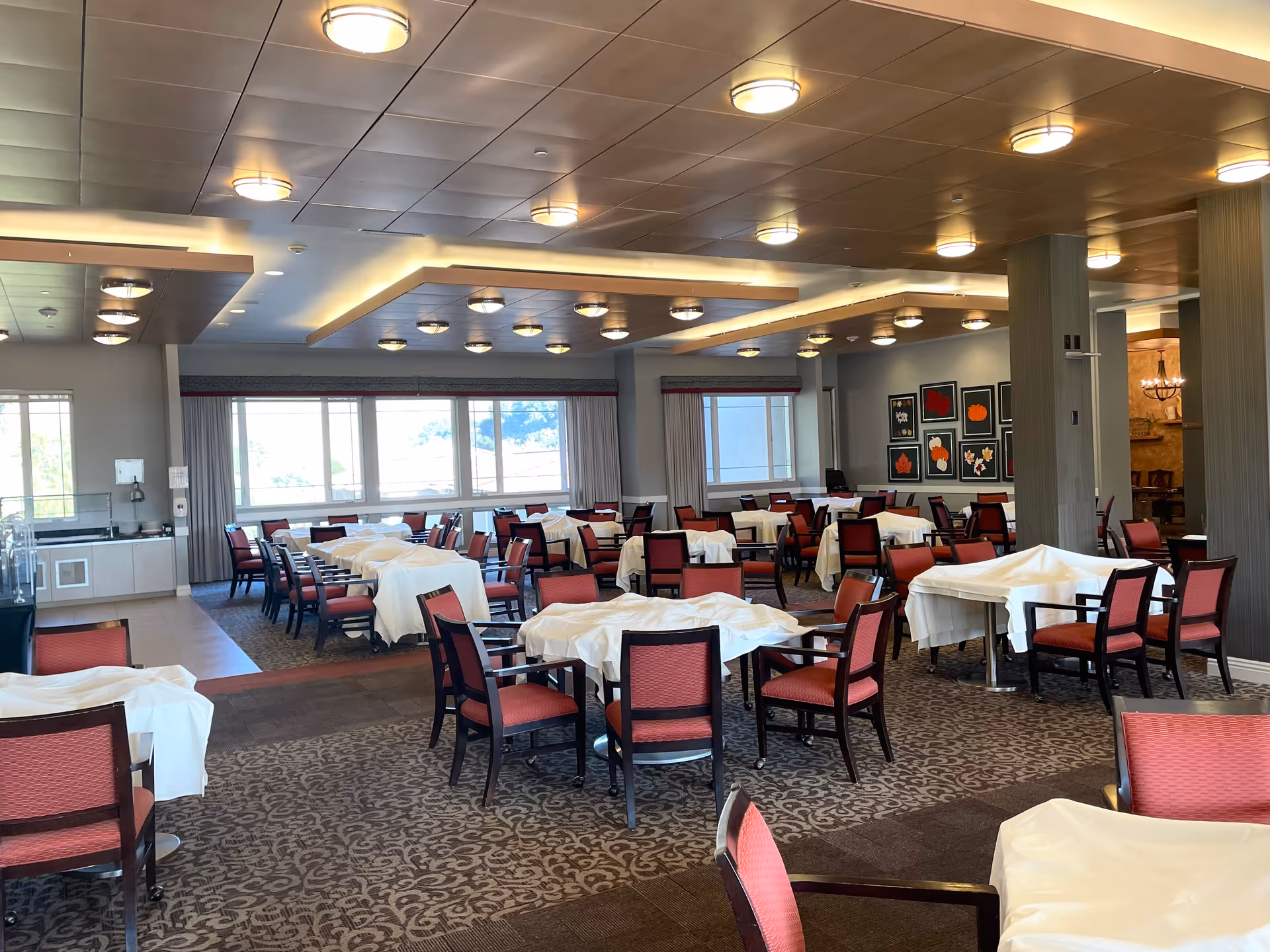 A spacious dining room with multiple tables covered with white tablecloths and surrounded by red cushioned chairs. The room has large windows letting in natural light, a patterned carpet, and modern ceiling lights. There are framed artworks on one wall and a small kitchenette area visible in the background.