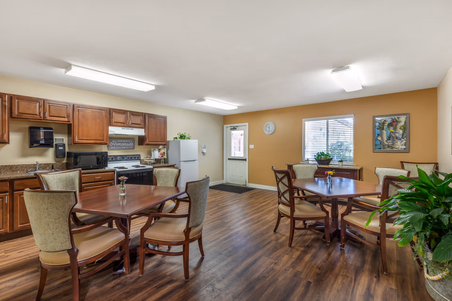 A communal kitchen and dining area with wooden floors, two wooden dining tables each surrounded by four upholstered chairs, wooden cabinets, a white refrigerator, a stove with a microwave on the counter, a wall clock, a window with blinds, and decorative plants and artwork on the walls.