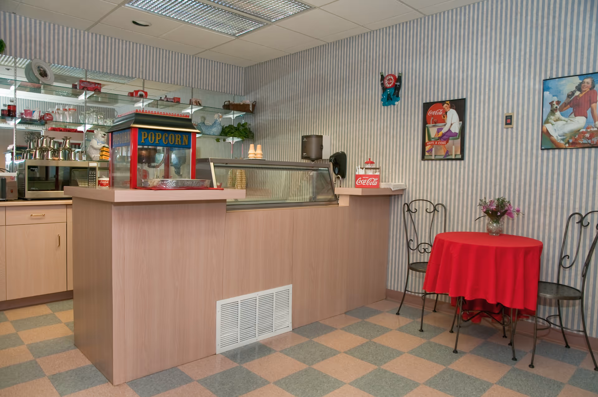 Small indoor snack counter with a popcorn machine, glass display case, and a red-tableclothed table with chairs.