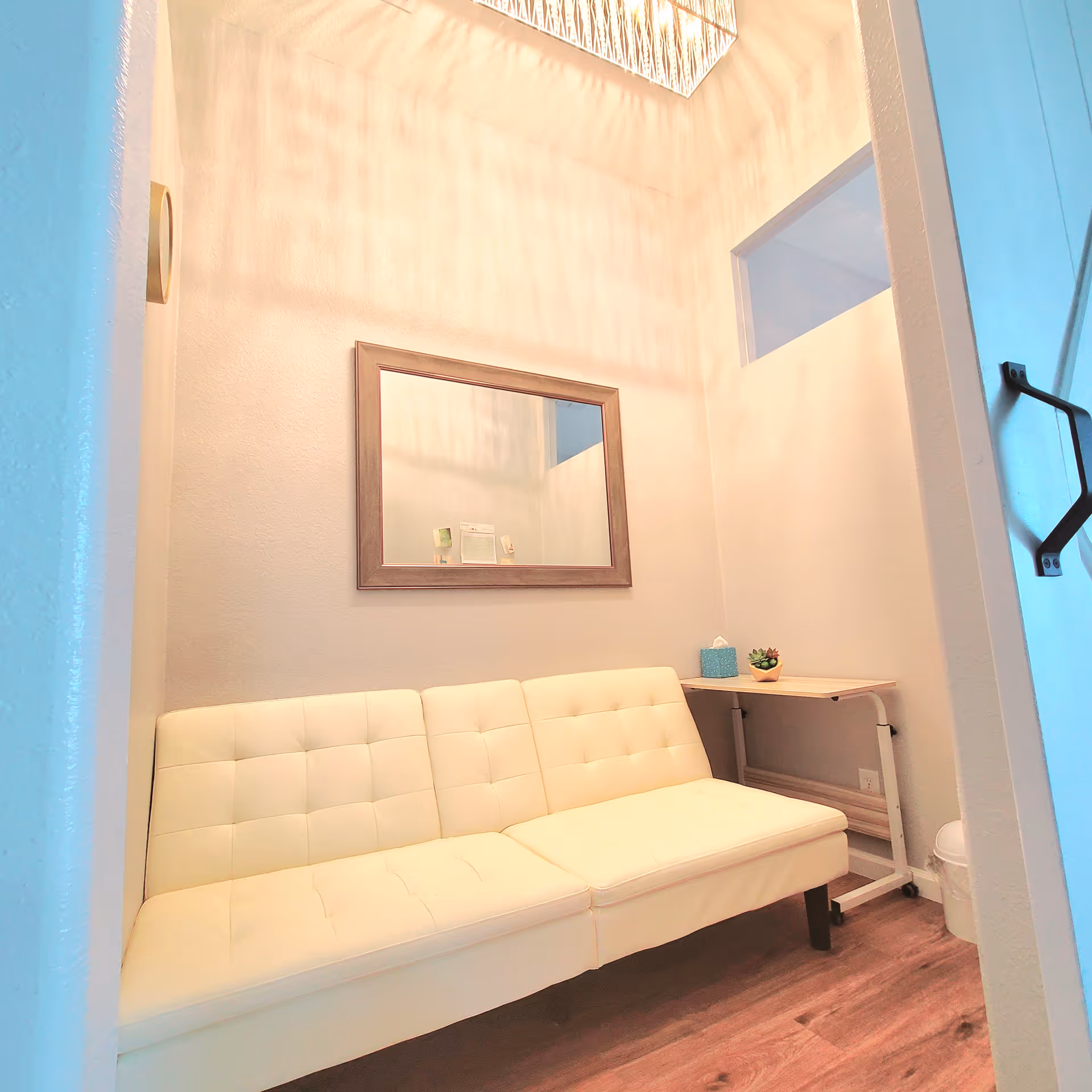 A small, cozy room with a white cushioned bench against a light-colored wall. Above the bench is a rectangular mirror with a wooden frame. To the right of the bench is a small wooden table with a tissue box and a small potted plant. The room has wooden flooring and a modern light fixture on the ceiling casting patterned shadows on the walls.