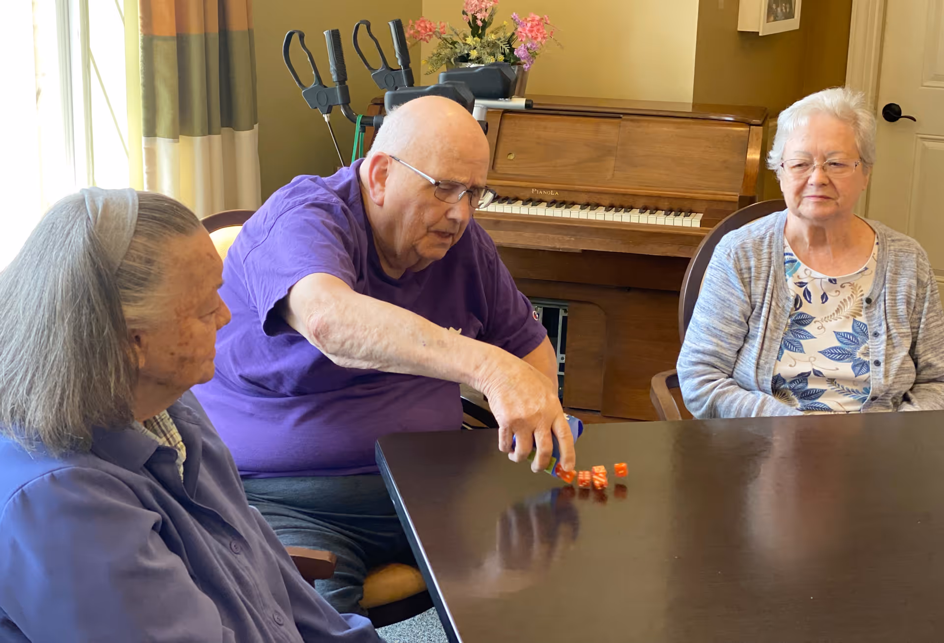 Three elderly individuals sitting around a table in a room with a piano in the background. One man in a purple shirt is rolling orange dice on the table, while two women watch. The room has a window with curtains and some flowers on top of the piano.