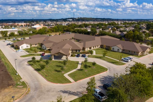 Aerial view of Legend Oaks Healthcare and Rehabilitation facility in Fort Worth, showing multiple connected single-story buildings with brown roofs surrounded by green lawns, driveways, and parking areas. The surrounding neighborhood with houses and trees is visible in the background under a partly cloudy sky.