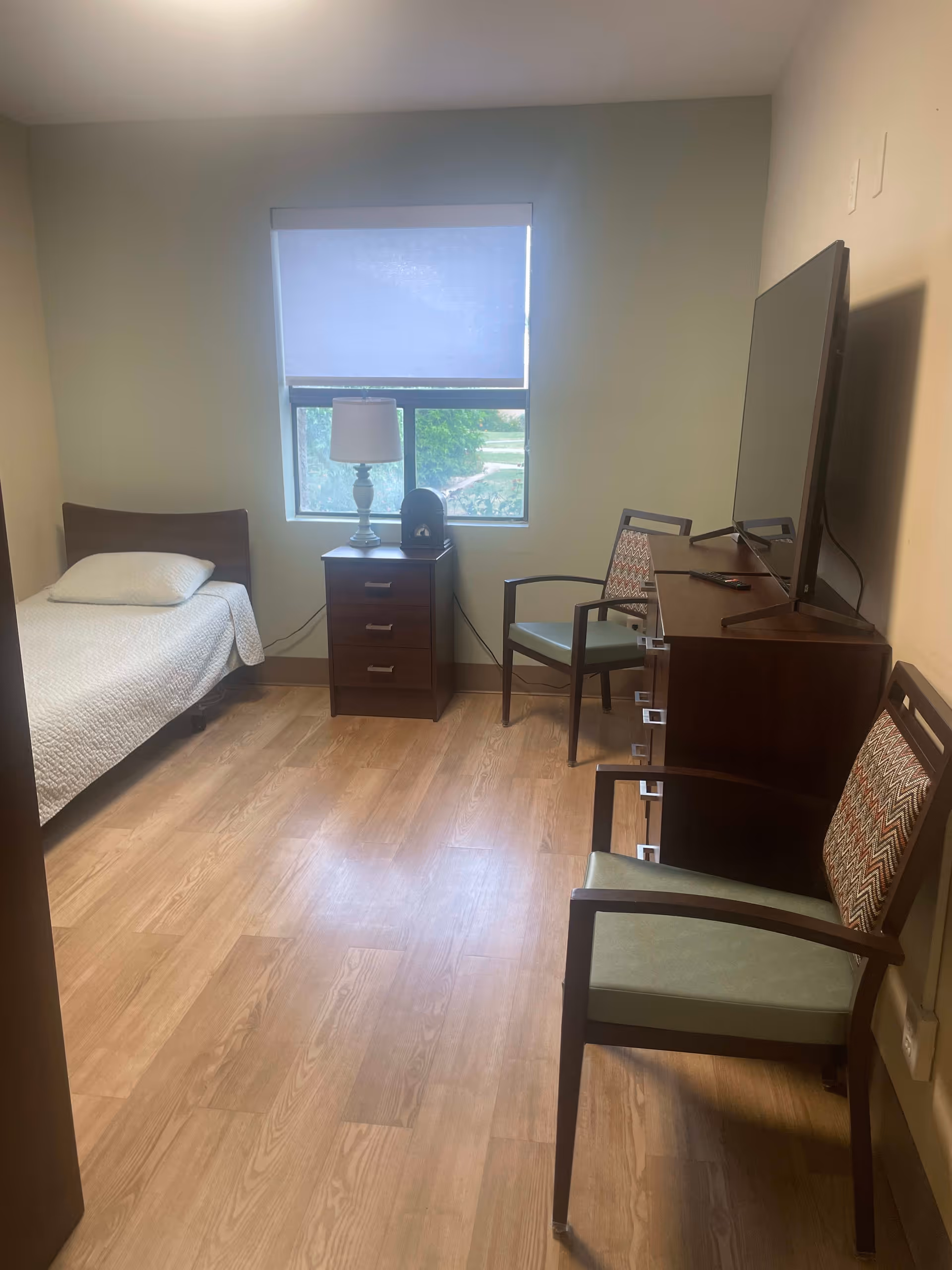 A small, simple bedroom with a single bed on the left side covered with a white quilt and a pillow. Next to the bed is a wooden nightstand with three drawers, a white table lamp, and a small decorative item. A window with a partially lowered white roller shade is above the nightstand. On the right side of the room, there is a wooden dresser with a flat-screen TV on top and two chairs with patterned backrests and green seats placed in front of it. The floor is light wood, and the walls are painted a light beige color.