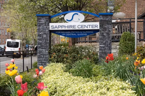 Outdoor view of the Sapphire Center for Rehabilitation and Nursing of Central Queens sign surrounded by colorful flowers and greenery, with a brick building and trees in the background.