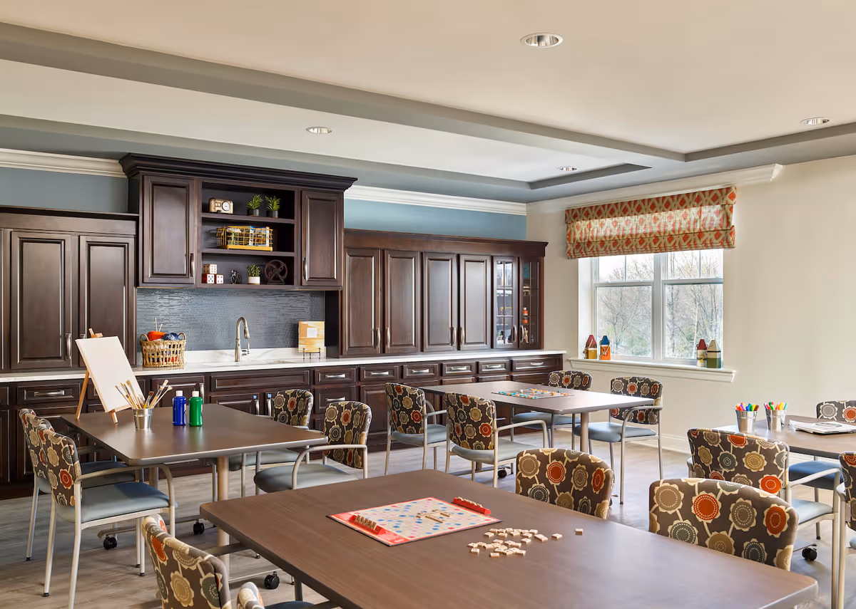 A bright activity room in an assisted living facility with several tables and chairs arranged for group activities. The tables have board games, art supplies, and paint bottles on them. The room features dark wood cabinets along one wall, a window with patterned curtains, and a light-colored ceiling with recessed lighting.