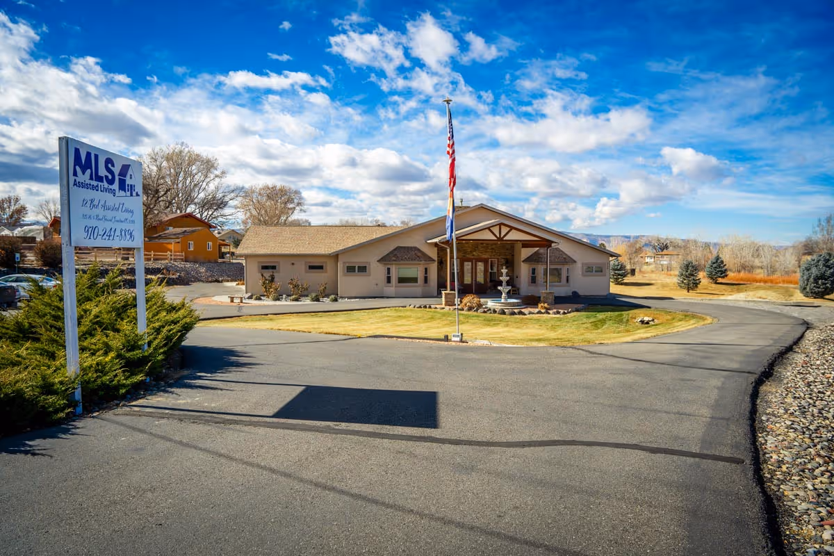 Exterior view of MLS Assisted Living CC LLC facility with a paved driveway leading to a single-story building. There is a sign on the left side displaying the facility's name and contact number. An American flag and another flag are positioned in front of the building, which is surrounded by grass, shrubs, and trees under a partly cloudy blue sky.