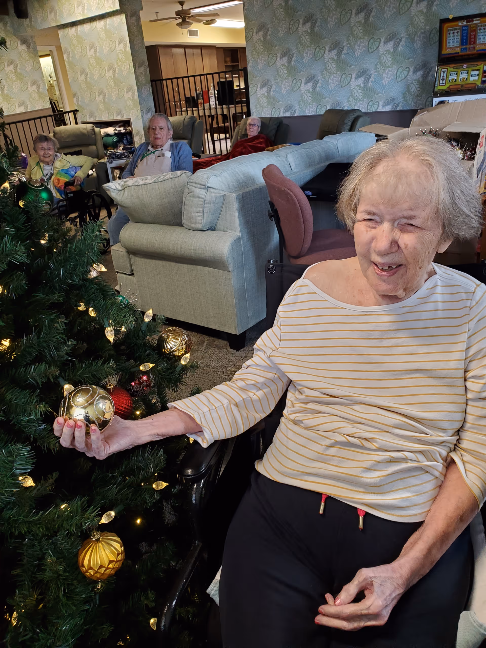 An elderly woman in a wheelchair smiling and holding a gold Christmas ornament near a decorated Christmas tree. In the background, three other elderly people are seated on couches in a cozy living room with patterned wallpaper and soft lighting.