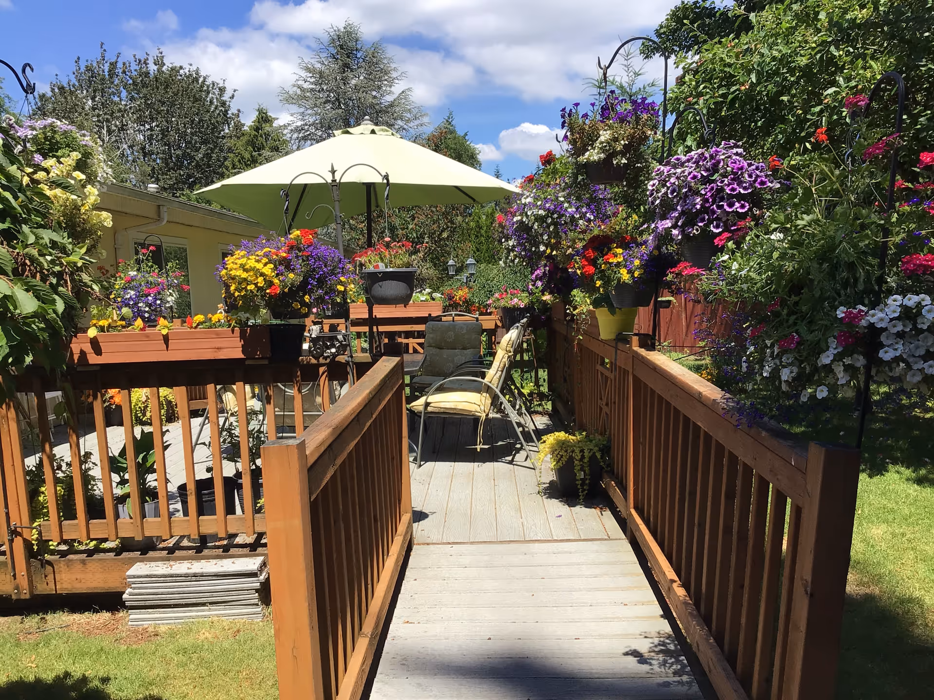 A wooden deck walkway lined with colorful hanging baskets and planters leading to a patio with chairs and a green umbrella in a sunny garden.