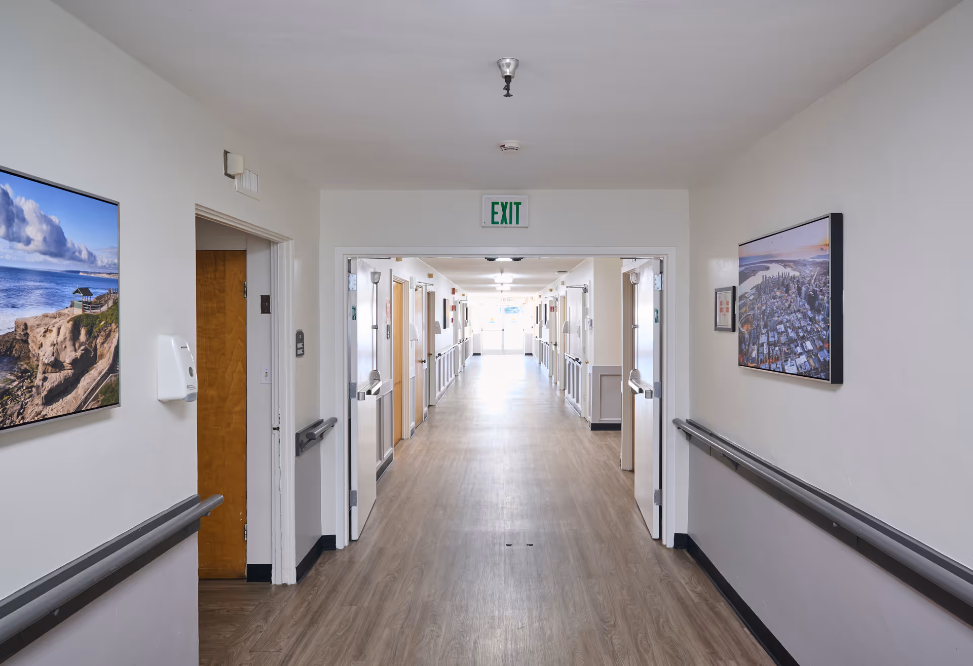 A long, well-lit hallway in a senior living facility with light-colored walls and wood flooring. There are handrails on both sides of the hallway, several doors leading to rooms, and framed landscape photographs hanging on the walls. An exit sign is visible above a doorway at the end of the hallway.