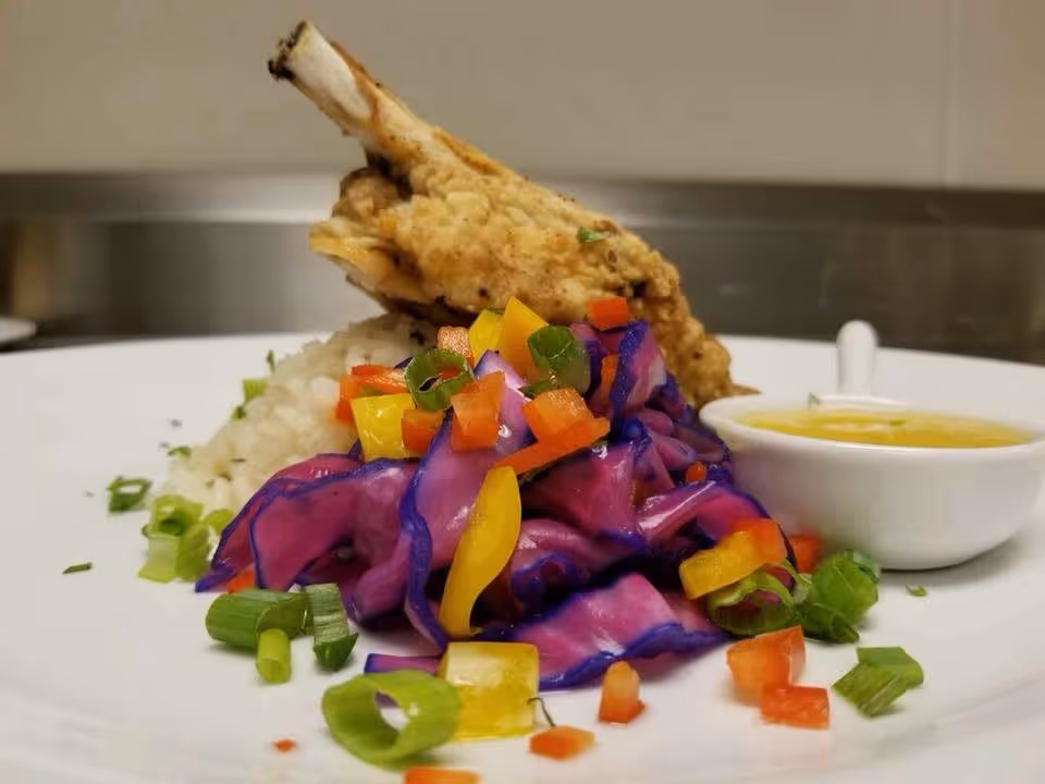 A plated meal featuring a piece of fried chicken, white rice, colorful sautéed vegetables including purple cabbage and diced red and yellow bell peppers, garnished with chopped green onions, and a small white bowl of yellow sauce on the side.