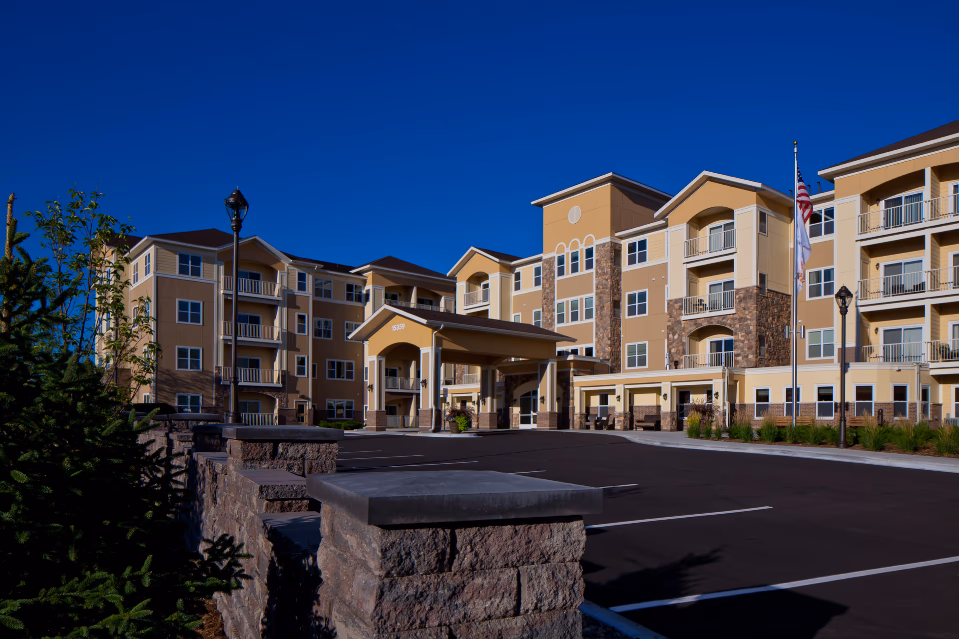Exterior view of a multi-story senior living facility building with beige and stone facade, balconies, and a covered entrance. There are flagpoles with flags and a paved parking area in front, under a clear blue sky.